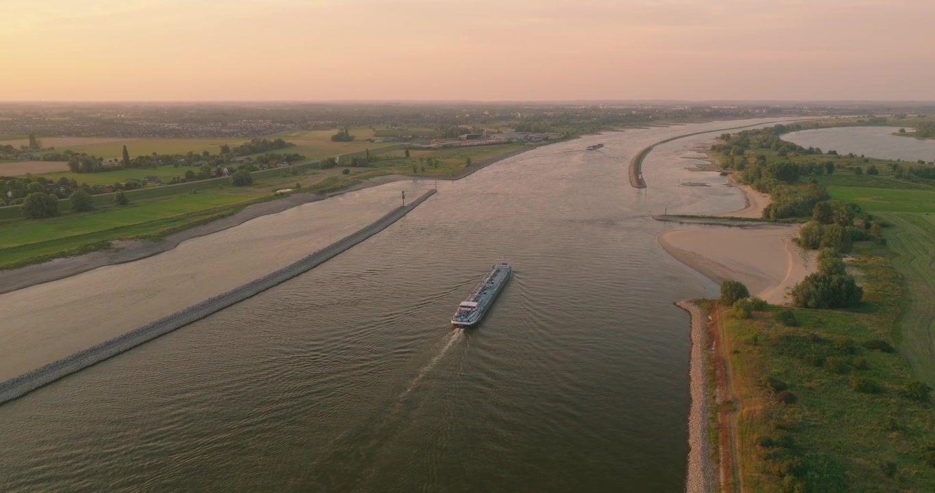 Spettacolare vista drone di una nave da carico che naviga lungo il fiume Waal al tramonto sotto un cielo arancione vibrante, con dighe, terreni agricoli rurali e rive alberate che creano un suggestivo paesaggio fluviale olandese