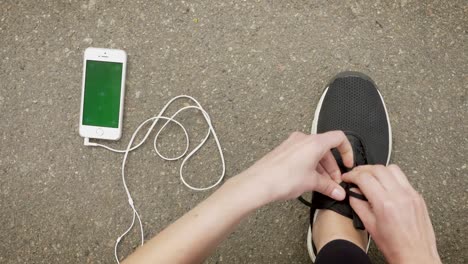 una chica deportista se ata los cordones de los zapatos mientras su teléfono inteligente y auriculares están en el suelo, pantalla cromada, concepto de comunicación