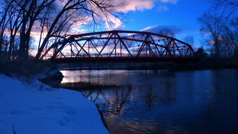 Lapso-De-Tiempo-De-Un-Pequeño-Puente-Rojo,-Arqueado,-De-Acero-Sobre-Un-Río-Al-Atardecer-Con-Reflejos-En-El-Agua-Y-Autos-Conduciendo-En-Un-Pequeño-Pueblo-De-América