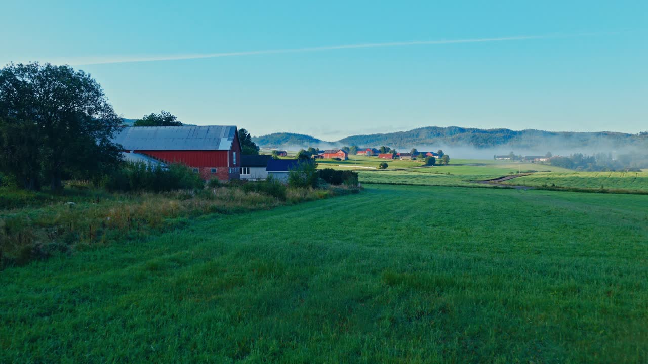 Rødt ladehus på grønne marker med bølgende bakker ved tåget solopgang. Luftdroneoptagelse.