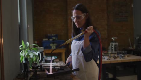 joyera caucásica en el taller con delantal y gafas, usando un quemador de gas