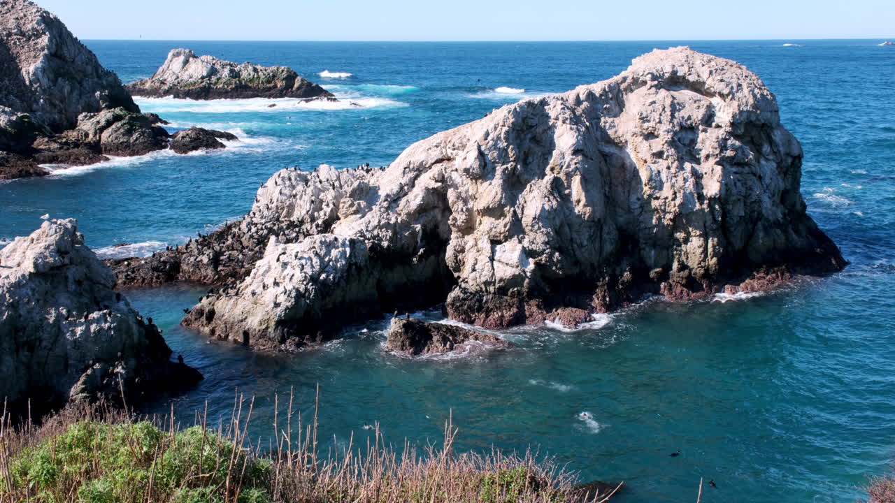 Large jagged California rock formations rising from the deep blue ocean, with waves gently breaking around their bases
