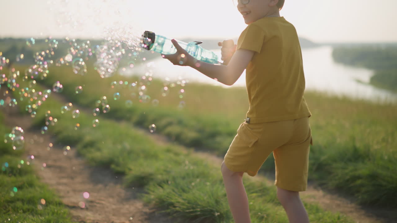 Young boy wearing a yellow shirt and shorts, with glasses, runs through a sunny grassy field while holding a plastic bubble gun that releases a stream of bubbles. The scene is set beside a serene lake