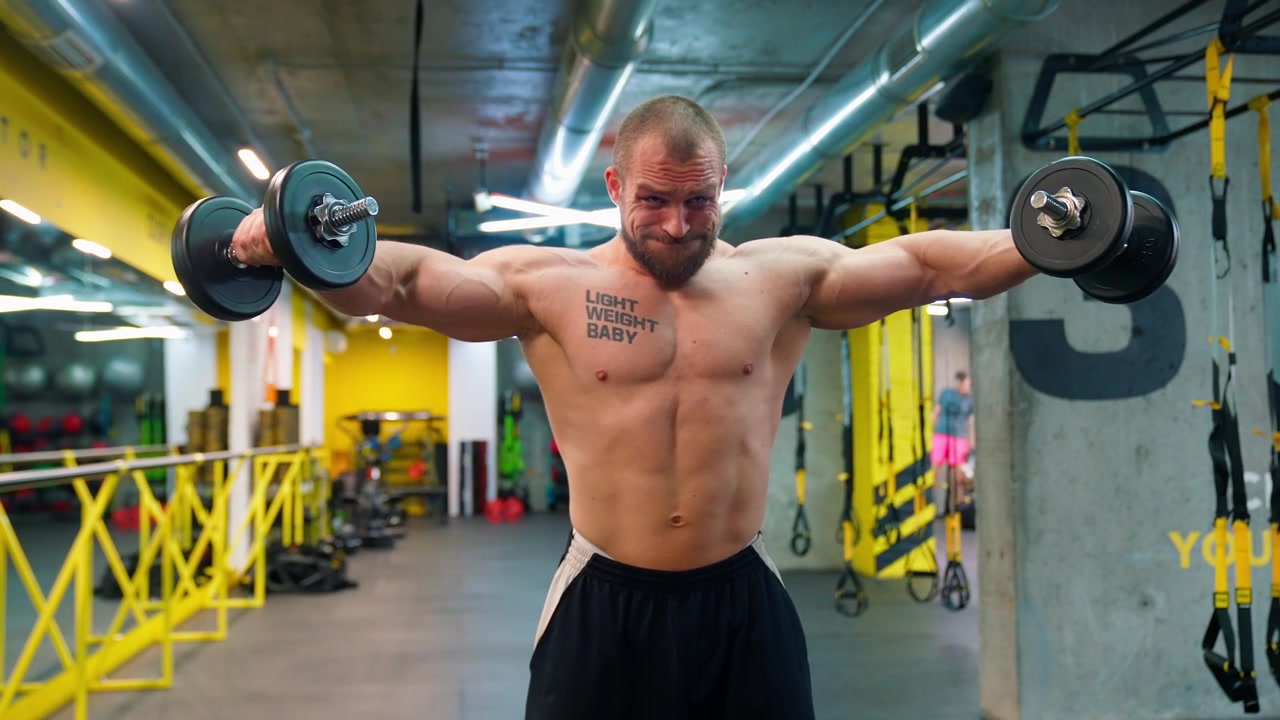 Homem musculoso sem camisa fazendo elevação lateral com halteres na academia