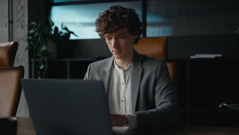 happy young guy with curly hair in a gray suit working and typing on a gray laptop while sitting at a table in a modern office