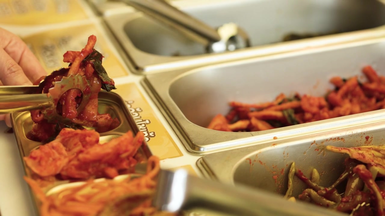 Primo piano di una mano che usa pinze metalliche per prendere kimchi da vassoi da buffet in acciaio inossidabile