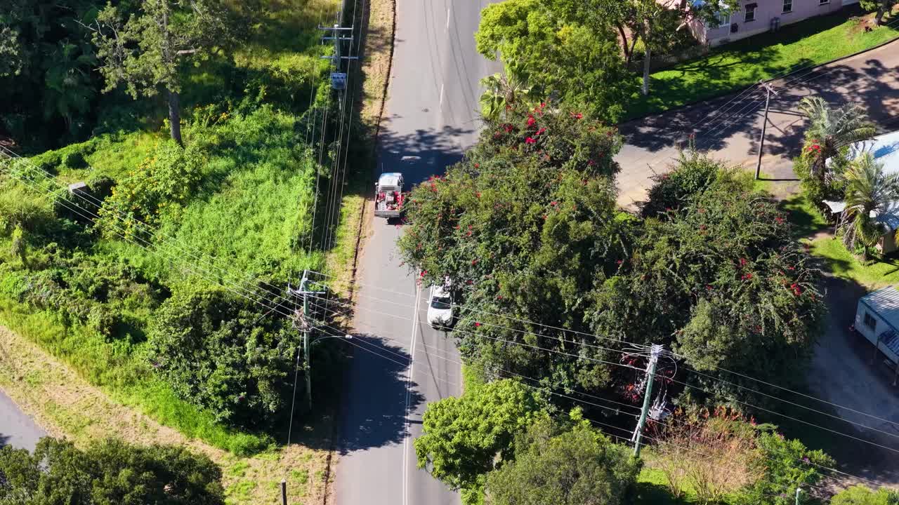Drone footage captures vehicles on a rural road surrounded by greenery and small buildings under bright daylight