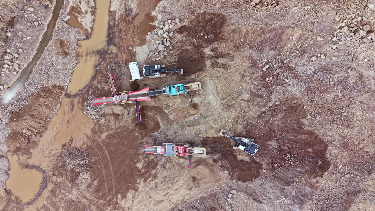 Construction equipment operates in a gravel pit in the afternoon near a stream while workers manage the site for building materials