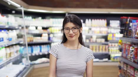 Porträt-Einer-Attraktiven-Jungen-Asiatischen-Frau-Mit-Einkaufswagen,-Die-Im-Supermarkt-Mit-Regalen-Mit-Milchprodukten-Im-Hintergrund-Steht,-In-Die-Kamera-Blickt-Und-Lächelt.-Handelsgeschäft-Und-Personenkonzept