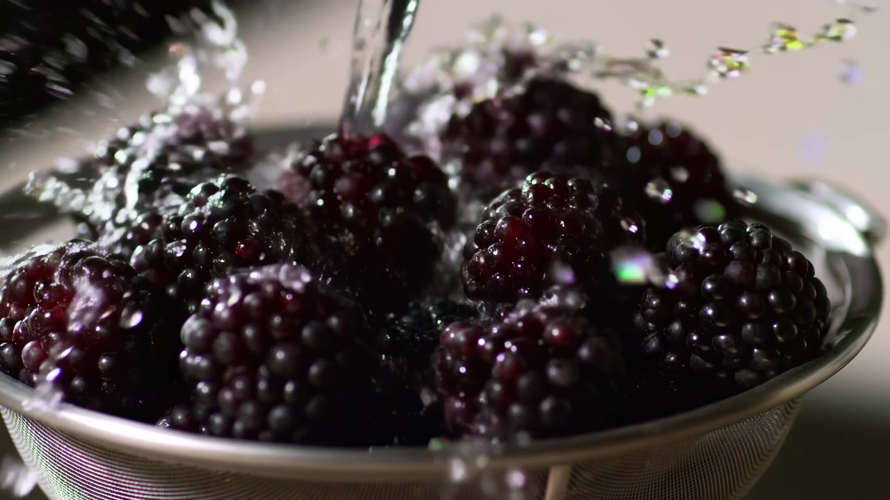 Primer plano de moras frescas en un colador de metal bajo agua corriente, centradas en el encuadre con iluminación lateral suave y gotas visibles en la fruta