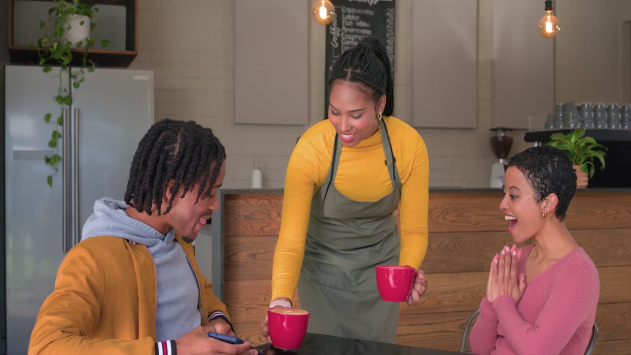 Diverse couple smiling as man showing smartphone screen and barista delivering red mugs for coffee