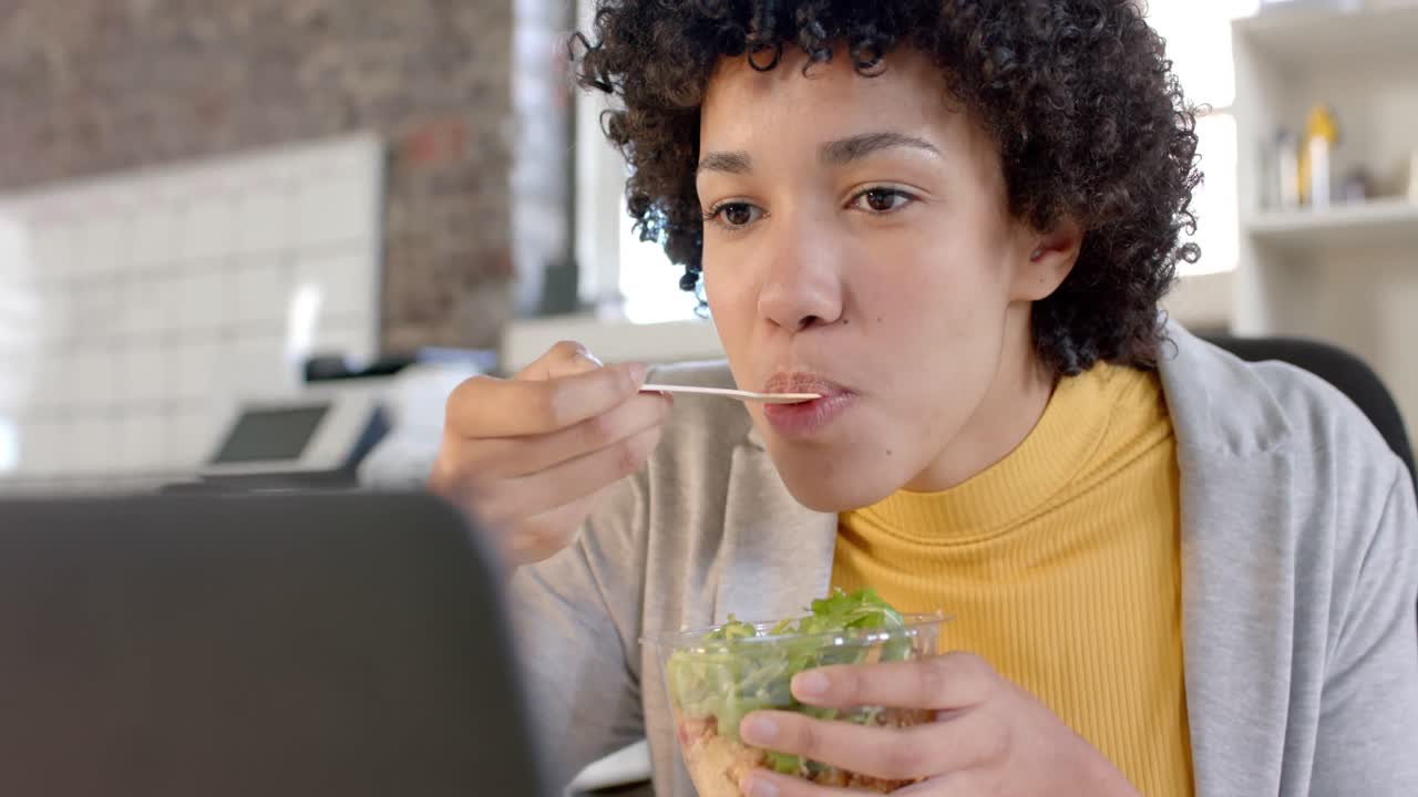 mujer de negocios casual biracial enfocada usando una computadora portátil y almorzando en la oficina en cámara lenta