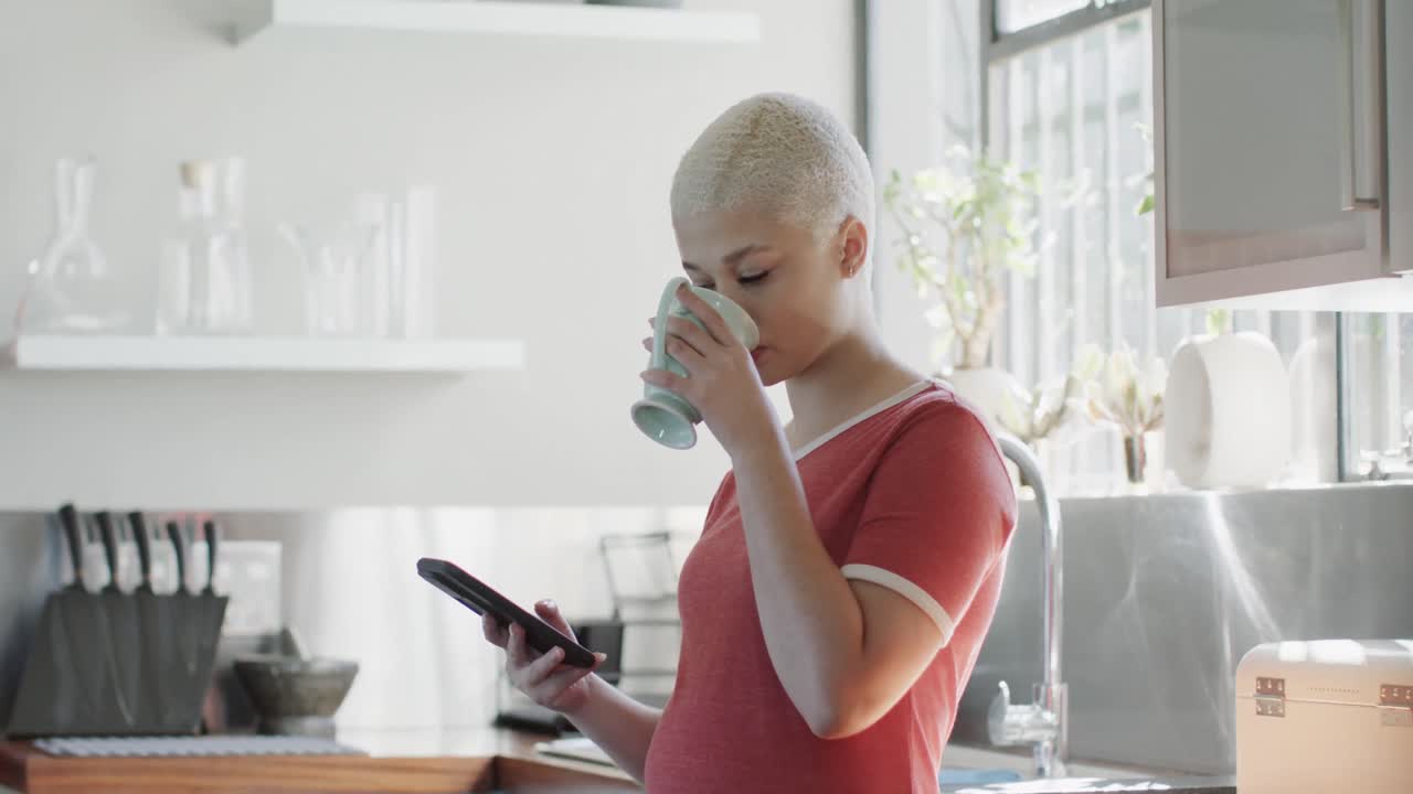 mujer biracial feliz bebiendo café y usando un teléfono inteligente en casa en cámara lenta