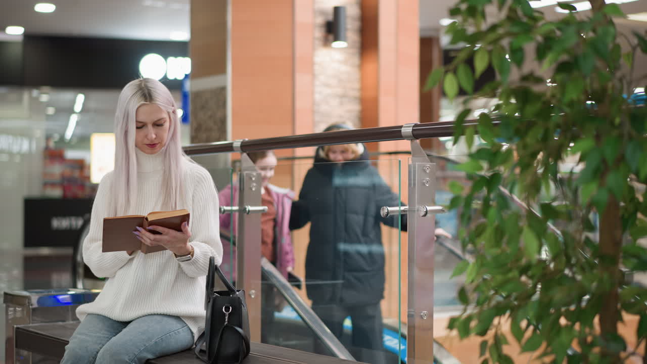 poised woman seated on bench flipping book pages with two hands, wearing cozy sweater, jeans and boots, black bag beside, mother and daughter entering moving walkway behind glass railing