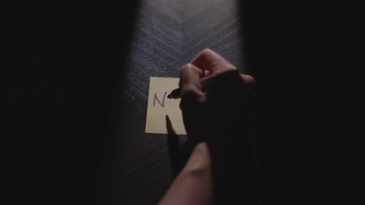 Slow motion close up footage of a hand writing out "November" on a paper note and placing it on a table. Shot in dramatic lighting for an impressive title or transition