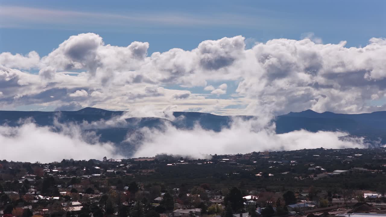 Drone performs a dynamic aerial ascent and track right over a southwestern town. Dramatic cumulus cloud structures frame the valley while atmospheric mist rises from the distant mountains