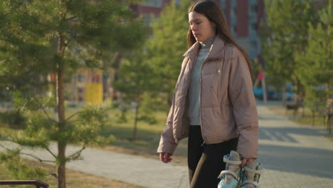 a young girl is walking through a sunlit park, holding a pair of rollerblades in one hand. she is dressed in a peach jacket, grey inner shirt, and black trousers