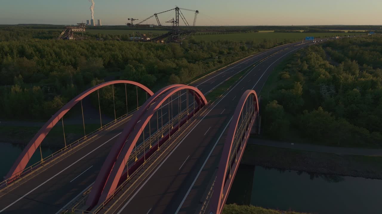 Aerial view of a highway bridge near Leipzig during sunset, with a massive coal excavator visible in the background. The scene blends modern roads, industry, and natural landscape.