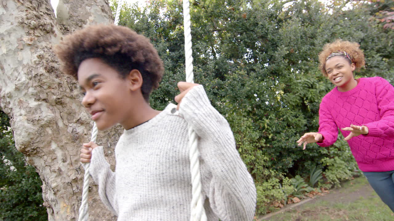 Happy african american mother pushing her son on tree swing in garden, slow motion