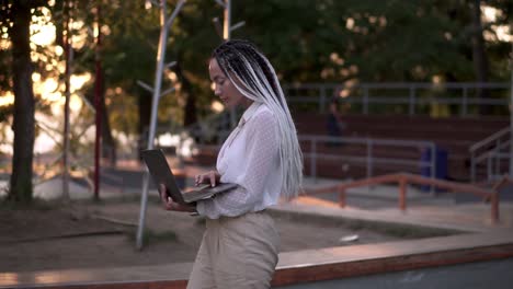 mujer encantadora con rastas de pelo blanco y negro caminando por la calle con una computadora portátil plateada abierta, escribiendo. bengalas de lentes y árboles