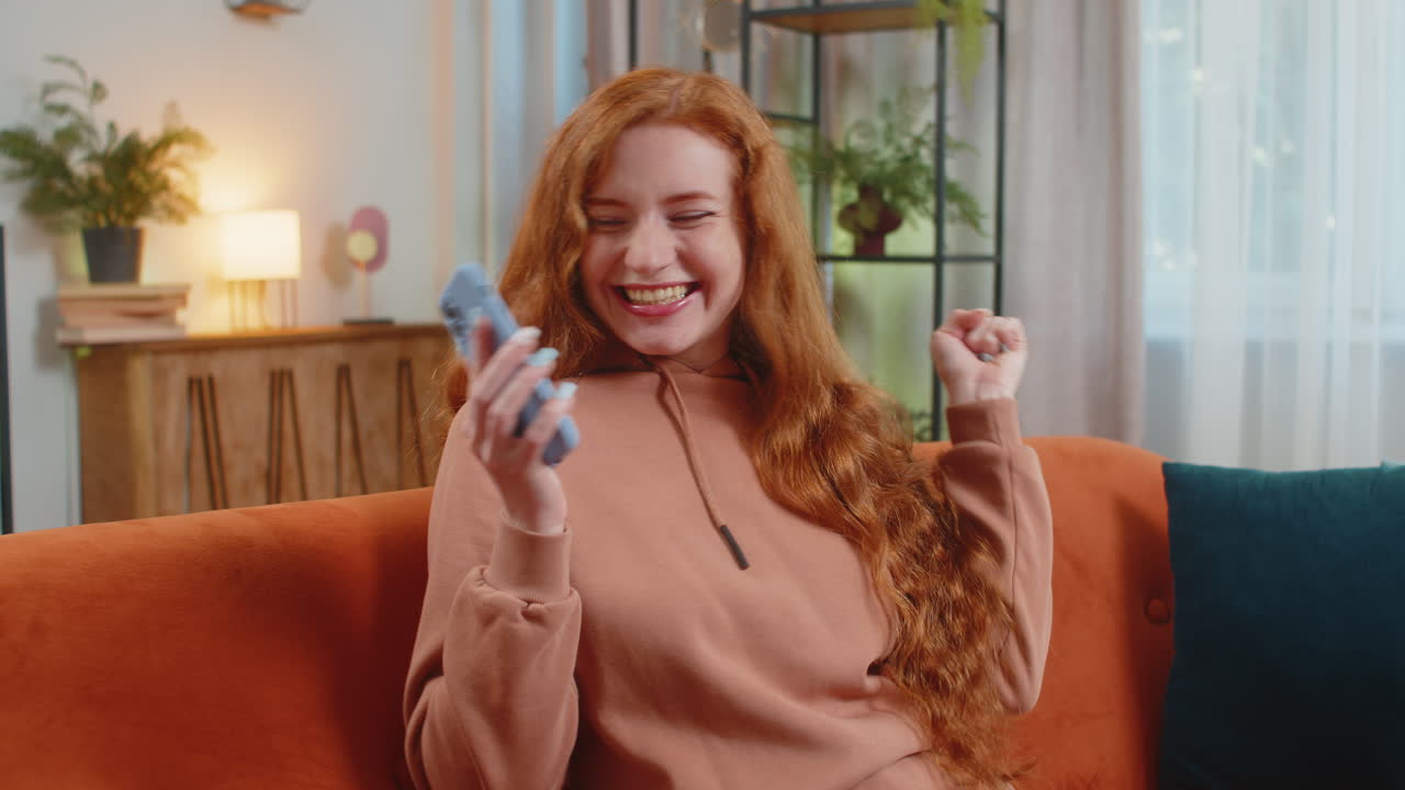 Happy young woman using cellphone celebrating success victory winning lottery on sofa in living room