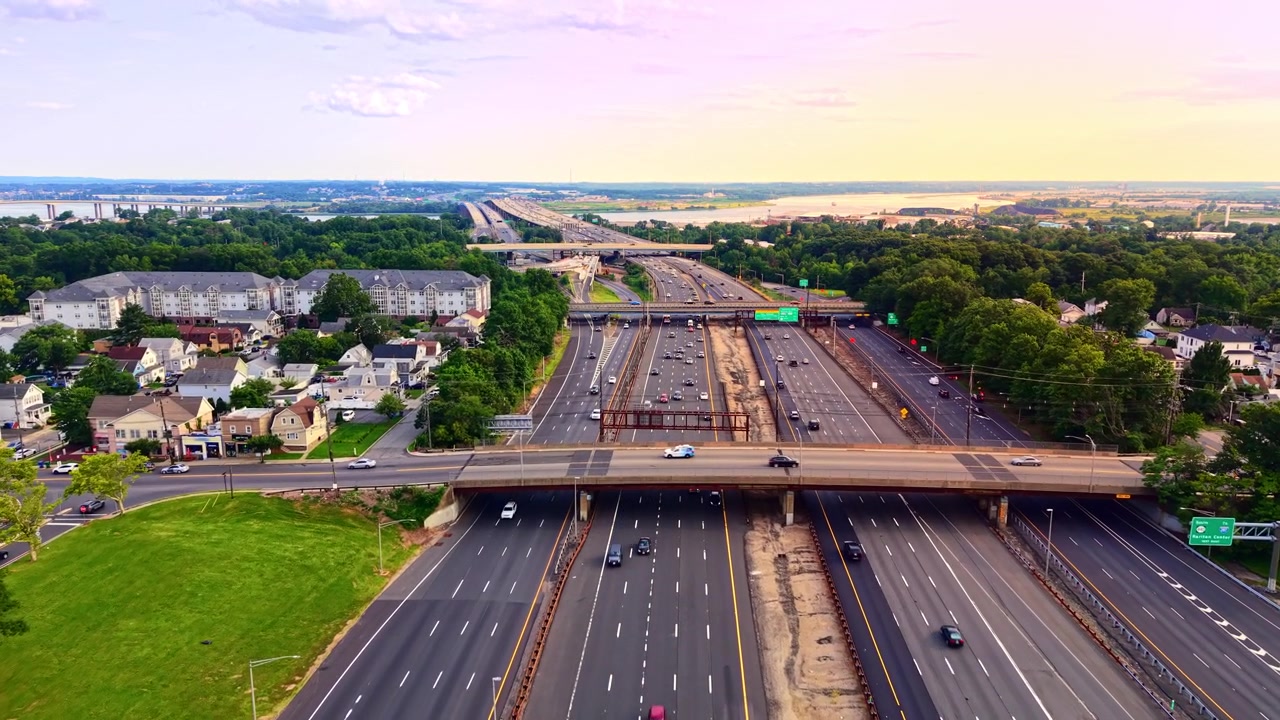 Wide aerial view of freeway overpass at sunset, New Jersey. Residential neighborhood, distant Governor Alfred E Driscoll bridge and traffic under pastel sky. Garden state parkway