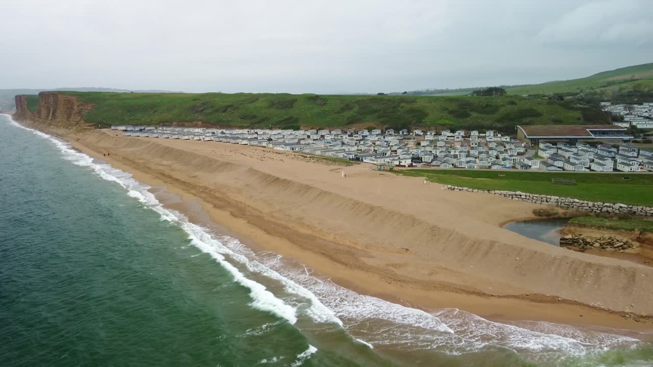 A campsite location on the coastline of the Jurassic Coast, Dorset. This aerial shot shows the lodges nestled in-between a river inlet and a grass top cliff with sheer cliff face down to the beach.