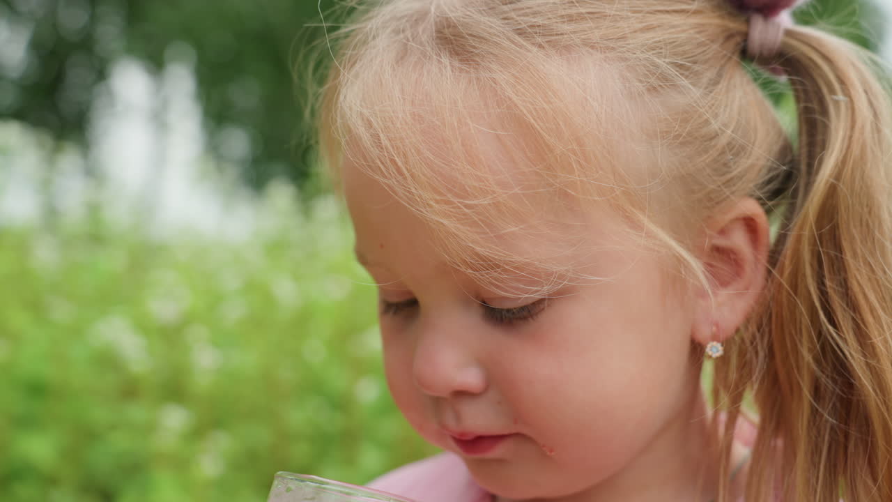 Primer plano de una niña pequeña bebiendo zumo al aire libre en una pradera soleada; una tímida niña pequeña prueba una bebida rosa con coletas y un pequeño pendiente, mirada suave entre sorbos, con un delicado fondo de hierba veraniega, reflejo lúdico