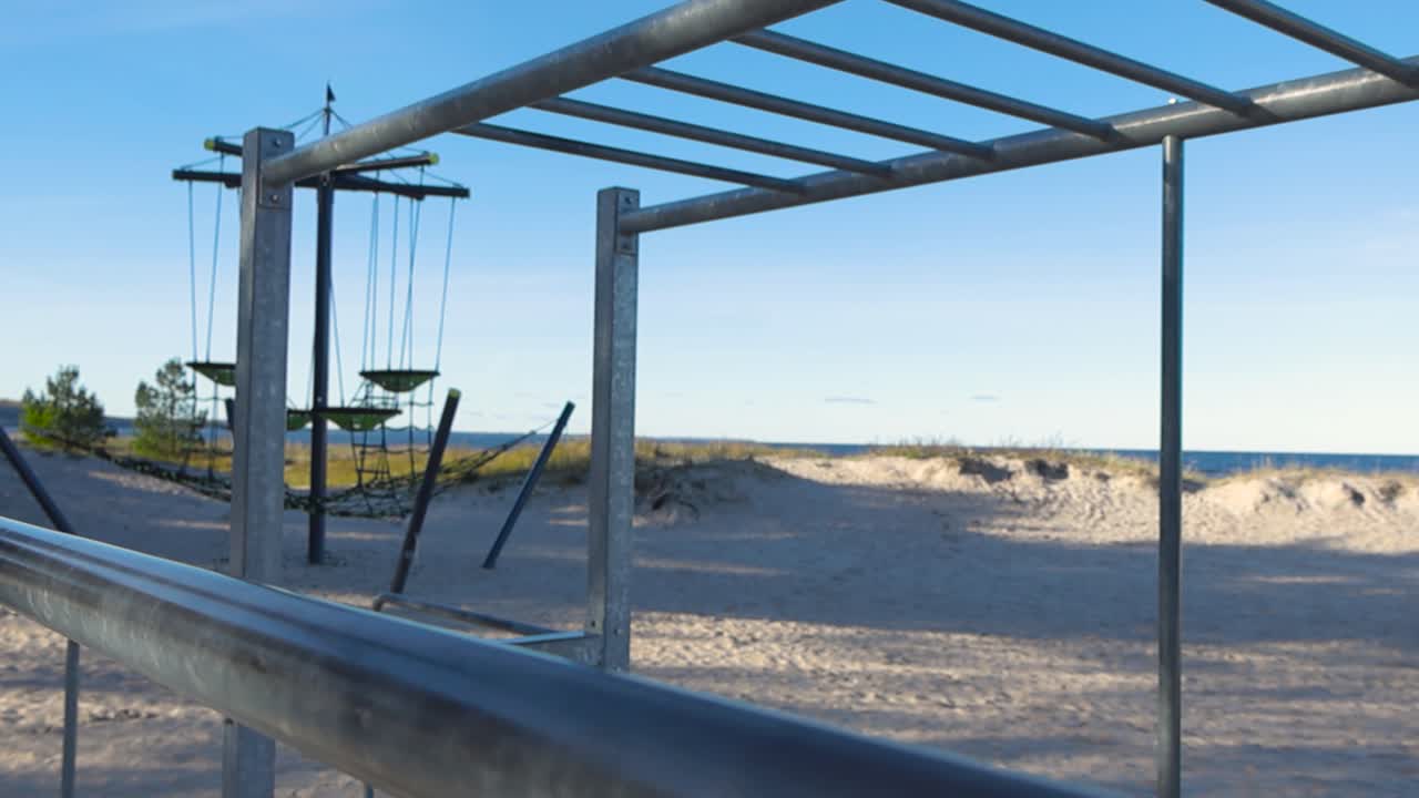 Outdoor gym and monkey bars at a beach and ocean is visible in the background. Sun is shining on the brown golden sand and some beach plants visible. Focus is changing from front to background.