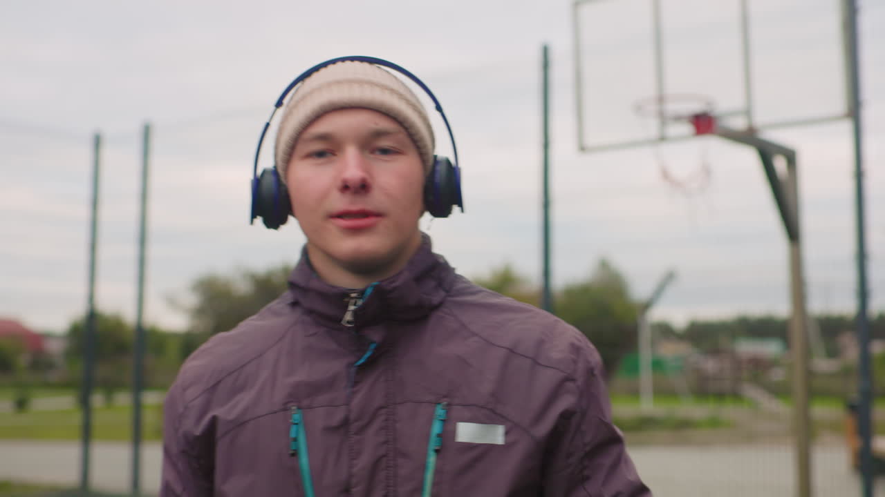 Jovem caucasiano em quadra de basquete, usando gorro e jaqueta, ouvindo fones de ouvido enquanto segura uma bola de basquete, céu nublado, parque suburbano, expressão pensativa, roupa casual urbana, treino lento