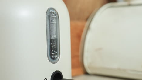 Close up of electric kettle with visible water level indicator showing steam and bubbling during heating process while red power light blinks in cozy kitchen setting