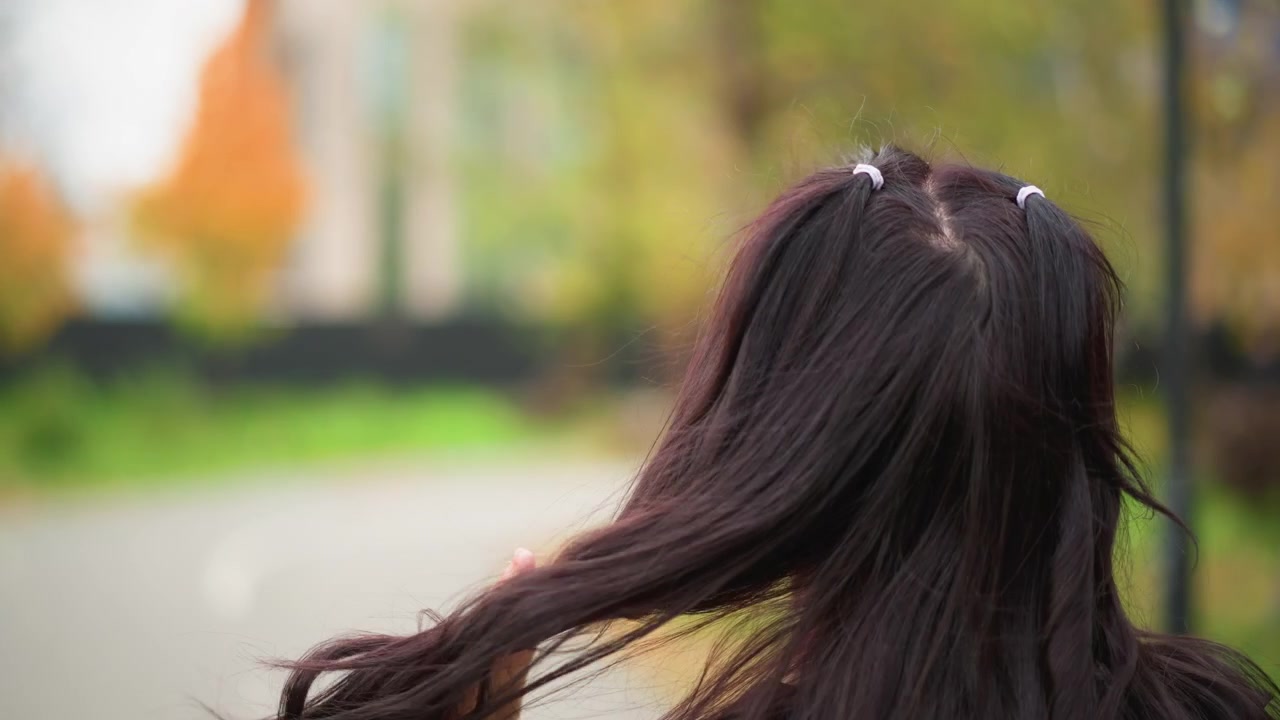 Close-up seorang wanita muda dengan rambut gelap panjang yang diikat dua kuncir kecil, berjalan di luar ruangan. Tangannya sesekali menyentuh wajahnya atau merapikan kuncirnya, menangkap gerakan-gerakan halus.