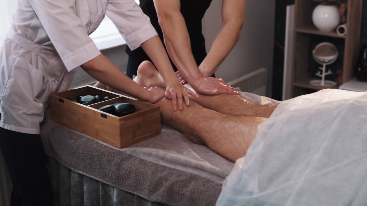 le mani di massaggiatori maschili e femminili stanno massaggiando le gambe di un maschio in abbigliamento sportivo usando la tecnica a quattro mani