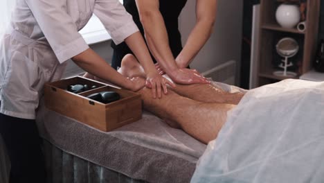 hands of male and female massage therapists are massaging the legs of a male in athletic clothing using the four-hand technique