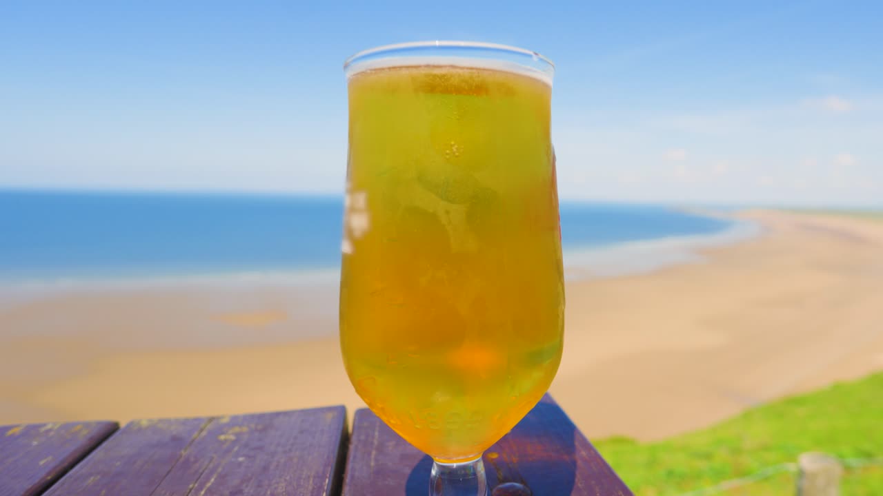 Refreshing Pint of Lager Beer on Picnic Table Overlooking Long Sandy Beach and Clear Blue Sea. Chilled Alcoholic Drink. Example of Holiday Vacation Rest and Relaxation.