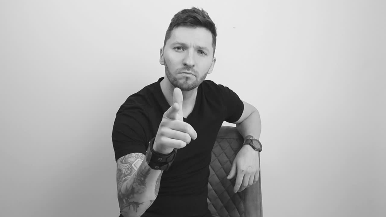 Grayscale footage of a tattooed man with short hair and beard lifting up his finger and pointing at the camera when nodding with approval. Man is sitting down in a chair and leaning against it.