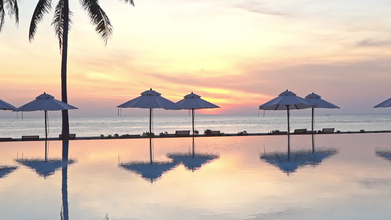 リゾートのスイミングプールの水面に映る、劇的なピンクとオレンジの夕焼けを背景にしたビーチパラソル。