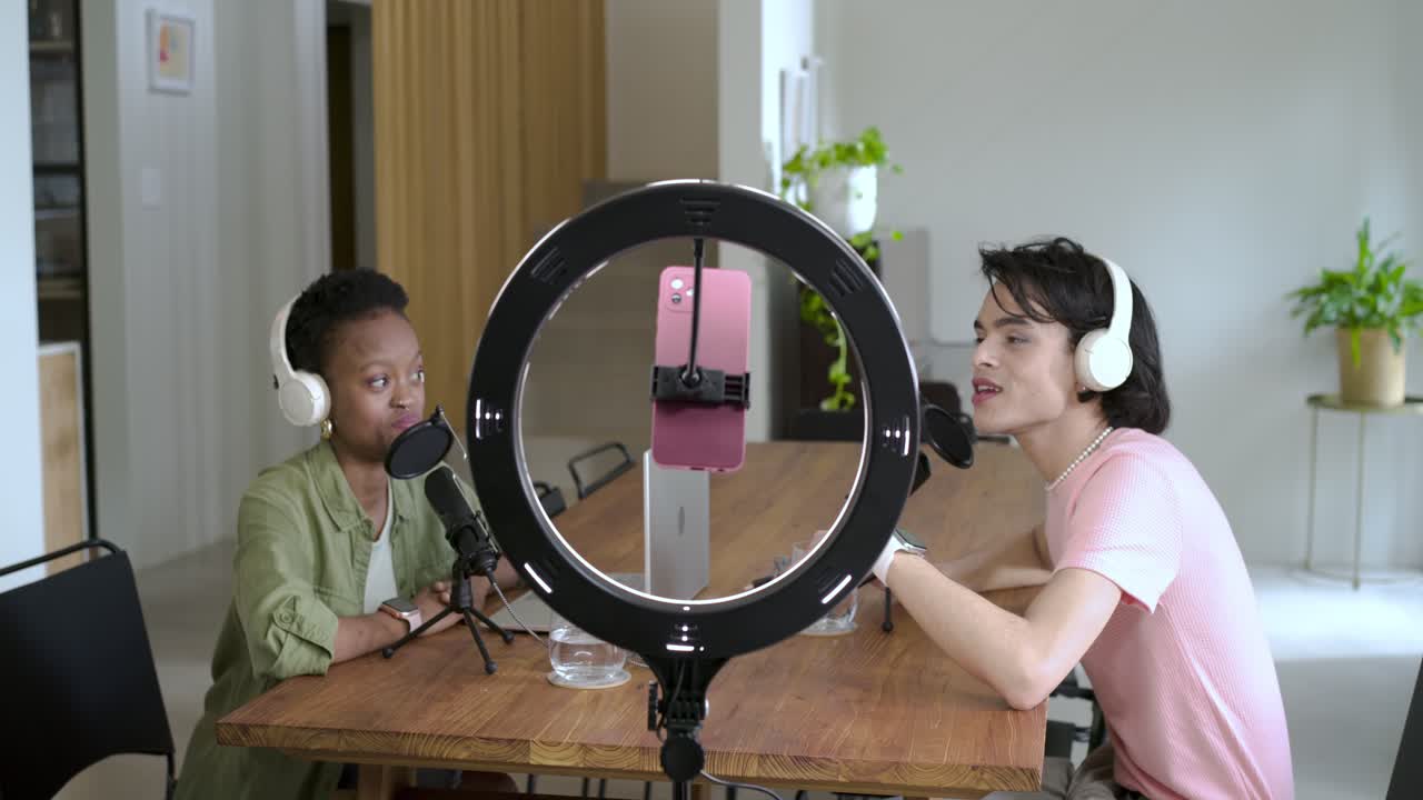 Diverse co-hosts leaning into mics discussing and gesturing at wooden table under ring light