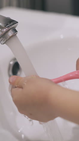 little girl cleans carefully pink toothbrush with water over white sink in light bathroom at home close view