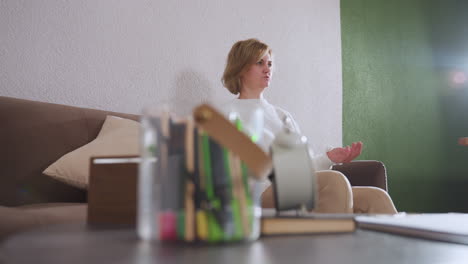 round view of healing seeker speaking hands gesturing during counseling session with therapist taking notes, cozy sofa with cushion, pen pot, clock on table, green and white wall background