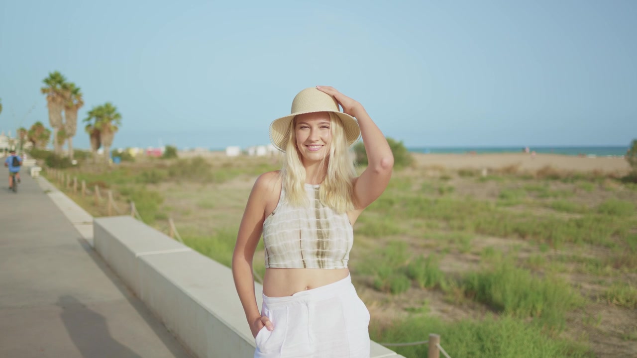 Giovane donna bionda felice con cappello che sorride in spiaggia.