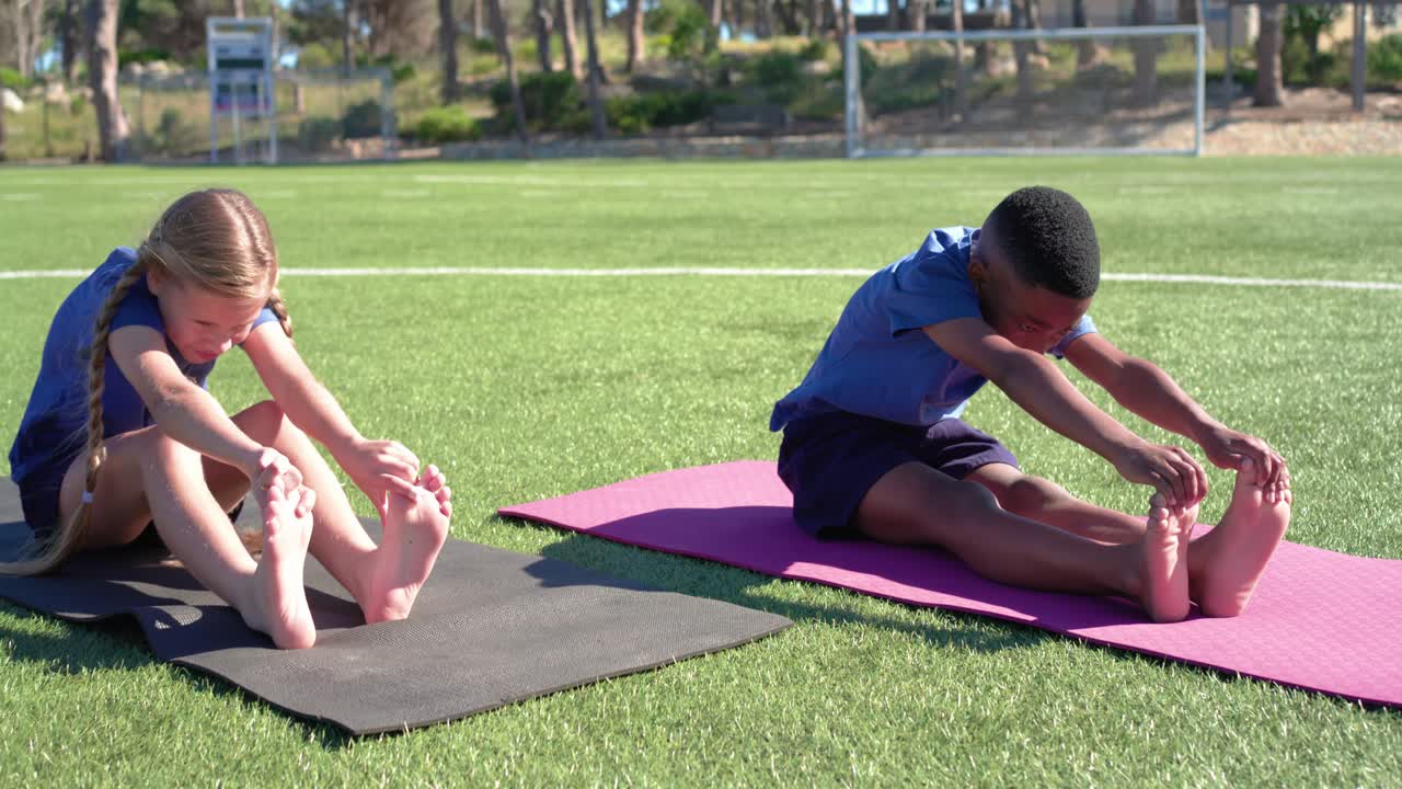 Diverse teenage classmates following coach cue, stretching forward on mats on turf near soccer goal