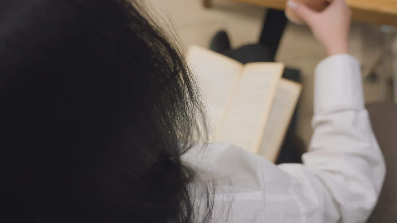 Close up rear view of woman with long dark hair wearing white shirt reading open book while seated, as she reaches forward and picks up juice cup placed on wooden table in calm indoor setting