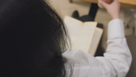 close up rear view of woman with long dark hair wearing white shirt reading open book while seated, as she reaches forward and picks up juice cup placed on wooden table in calm indoor setting