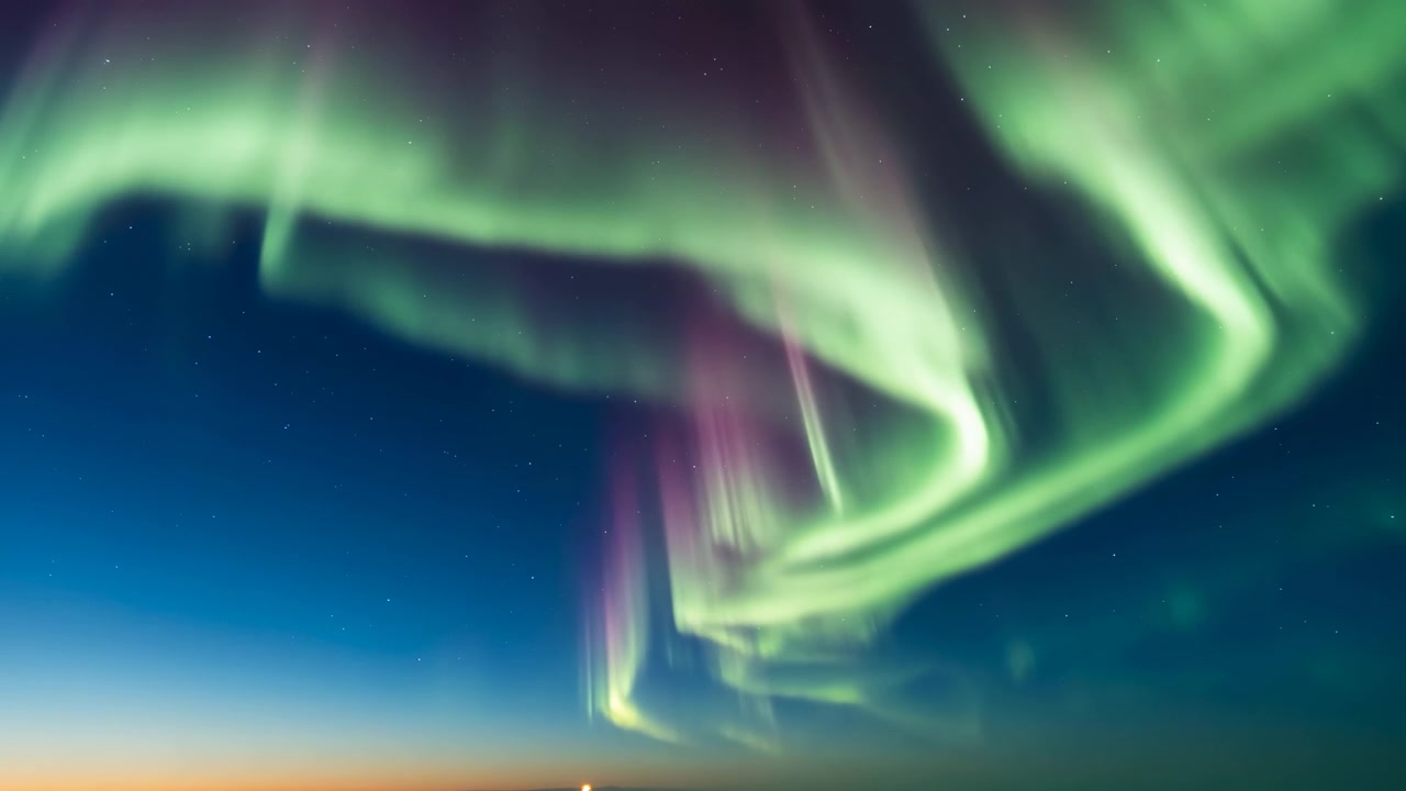 Ampia vista verso l'alto di un'aurora vivida che scorre attraverso un cielo indaco profondo, formando tende a strati, pilastri verticali, un bagliore soffuso e un contrasto radioso all'orizzonte in un'atmosfera notturna remota