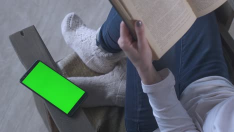 girl with short hair in jeans sits at home in armchair and reads book in winter. near mobile phone with green screen. woman with telephone. female cell phone. chroma key cellphone greenscreen.