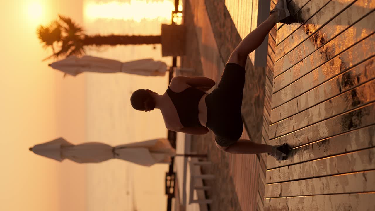 Vertical video: Brunette girl with tied hair in black sportswear doing lunges and stretches on a sunny beach during a golden sunrise in summer