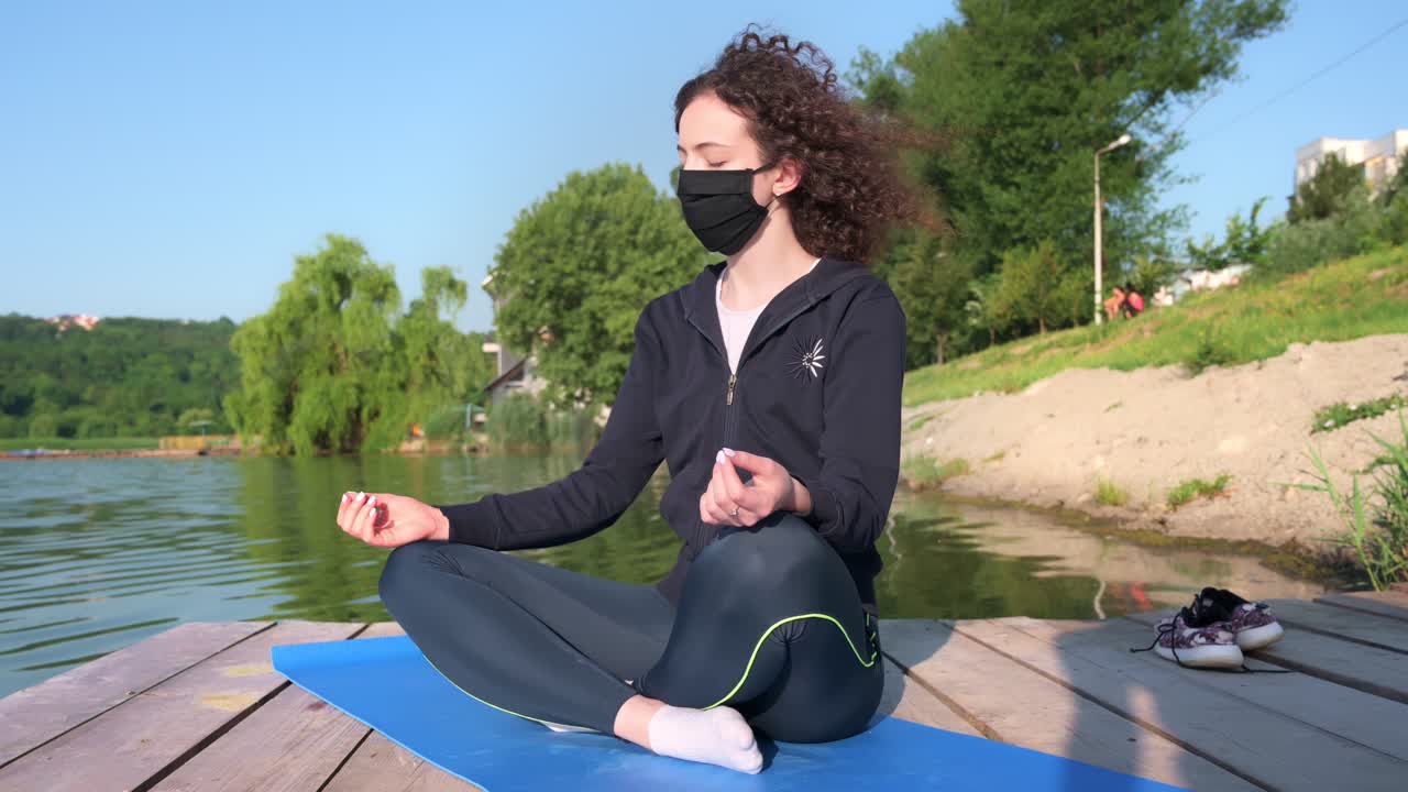 Young curly woman practicing yoga near the lake while wearing black protective mask. Sunny day. Meditating in lotus position on the yoga mat. Corona Virus idea. Chisinau, Moldova