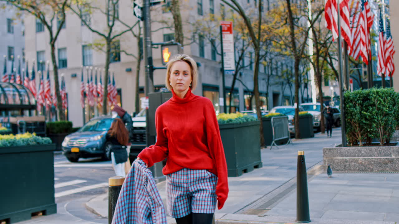 New York, USA, 5 ottobre 2025: Ritratto di donna con dolcevita rosso a Rockefeller Plaza. Ritratto di donna bionda con maglione dolcevita rosso in piedi vicino al cartello stradale e alle bandiere di Rockefeller Plaza