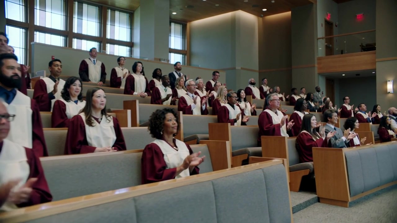 Coro che si alza in vesti bordeaux e stole bianche che applaude mentre inizia l'applauso in chiesa, telecamera che si sposta a destra
